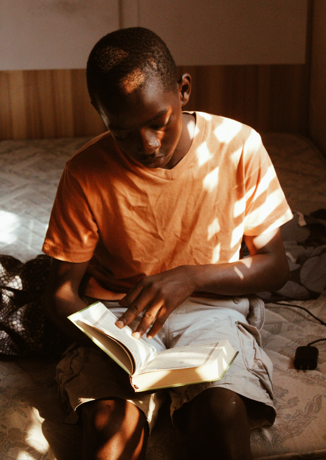 A boy reading a book while sitting on a bed.