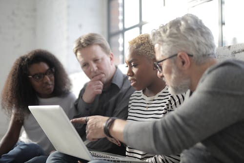 Free Middle aged employee in eyeglasses pointing at netbook screen with finger while sitting near multiracial pensive colleagues near window in afternoon Stock Photo