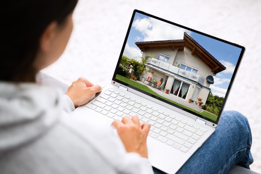 A woman on a couch with a laptop showing a house on the screen.